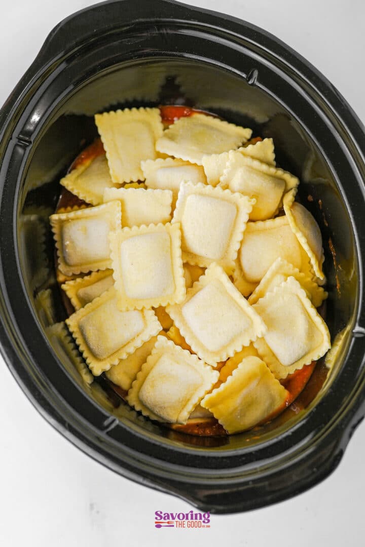 Uncooked ravioli arranged in a black slow cooker on top of a layer of tomato sauce, creating an easy lazy lasagna.