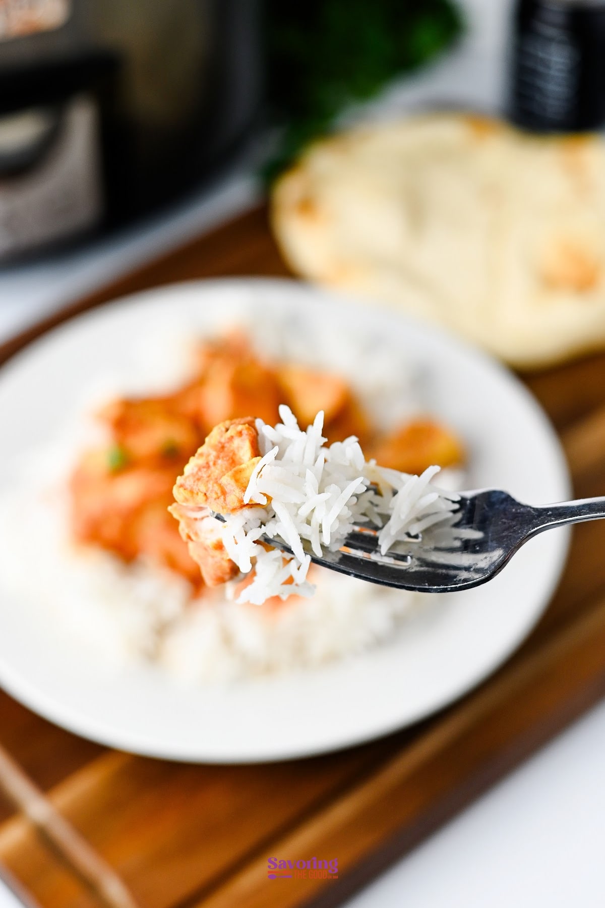 A fork holding a piece of slow cooker chicken tikka masala and white rice is in focus, with a plate of rice and curry and naan bread in the background.
