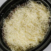 A slow cooker filled with lazy lasagna, featuring a layer of shredded cheese on top of savory ingredients, ready to be cooked.