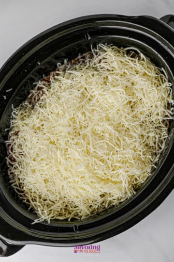 A slow cooker filled with lazy lasagna, featuring a layer of shredded cheese on top of savory ingredients, ready to be cooked.