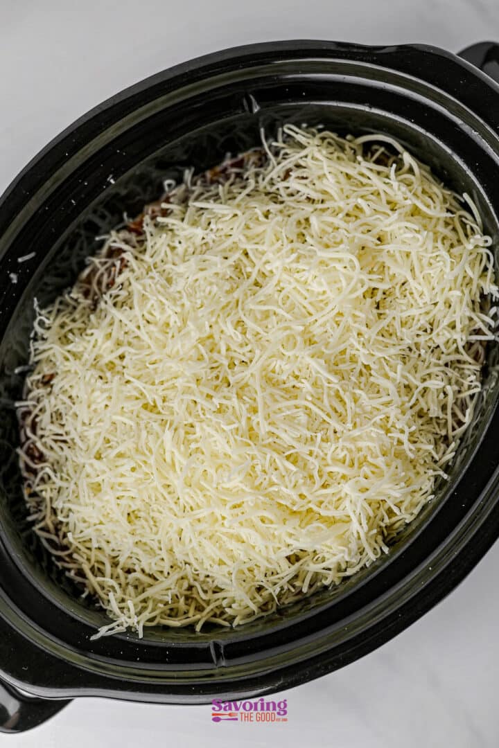 A slow cooker filled with lazy lasagna, featuring a layer of shredded cheese on top of savory ingredients, ready to be cooked.