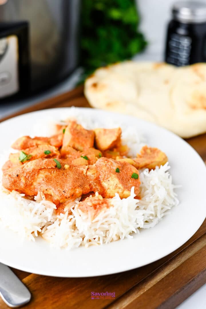 A white plate with white rice topped with pieces of slow cooker chicken tikka masala in an orange sauce, garnished with herbs, with naan bread in the background.