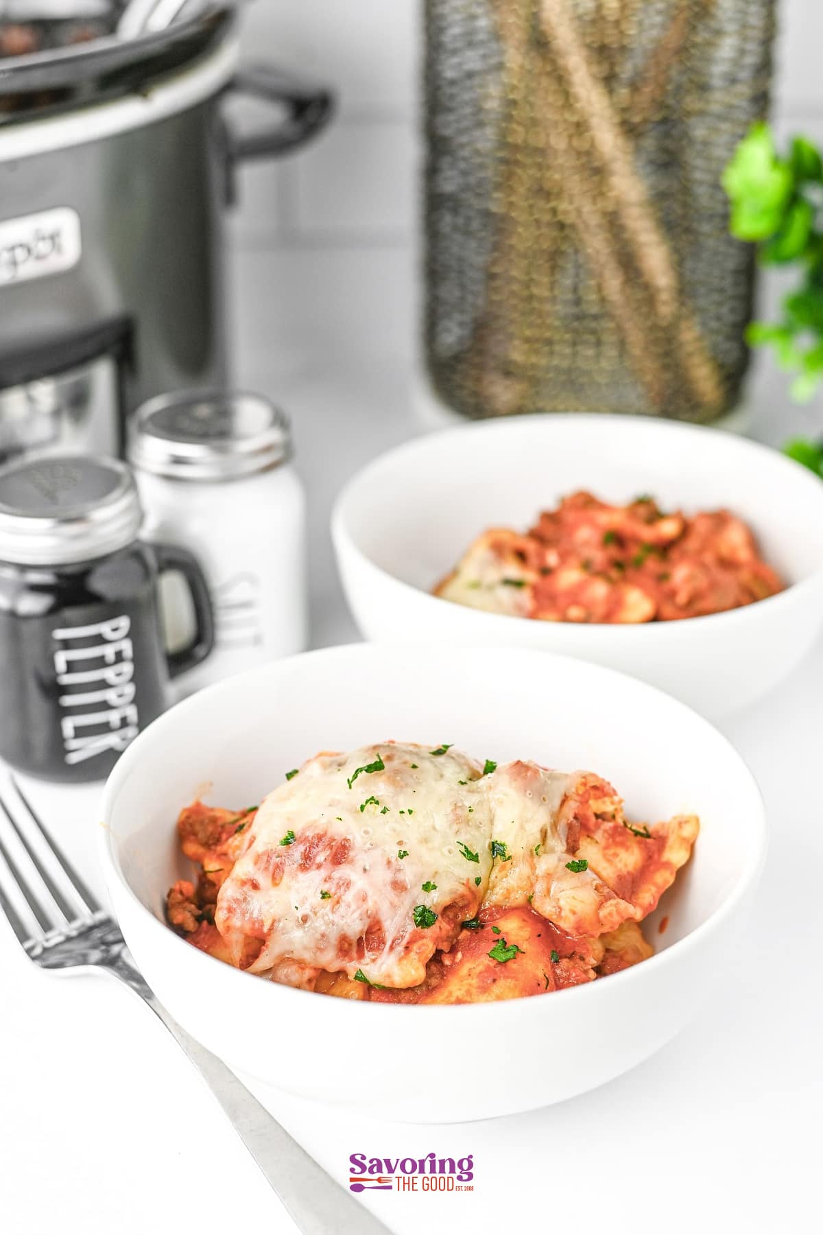 Two white bowls filled with cheesy pasta-this lazy lasagna is topped with herbs and sits near salt and pepper shakers on a white surface, with a slow cooker in the background.
