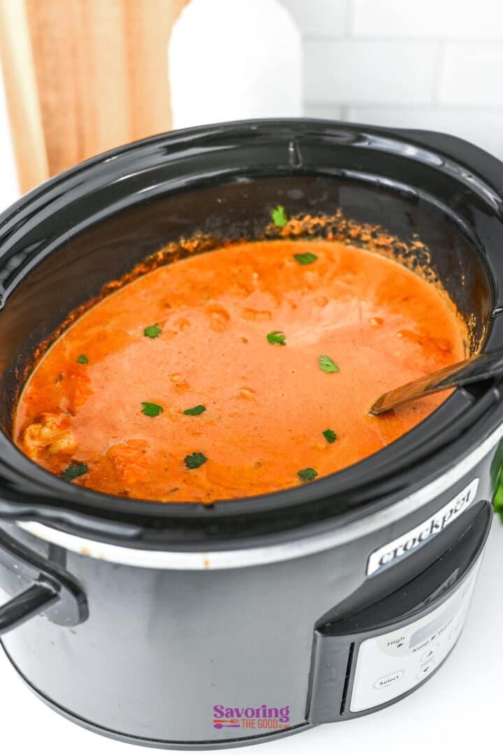 A slow cooker chicken tikka masala simmers in a crockpot, its creamy orange sauce garnished with small green herb pieces, with a spoon resting inside.