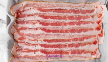 Several raw bacon strips are arranged side by side on a parchment-lined baking sheet. The image features the "Savoring the Good" watermark at the bottom.