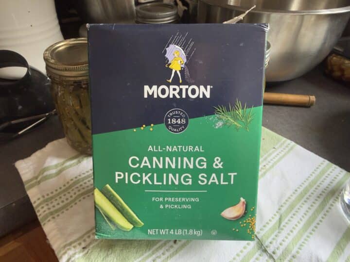 A box of Morton All-Natural Canning & Pickling Salt sits on a striped kitchen towel, with a jar of preserved vegetables and mixing bowls in the background.