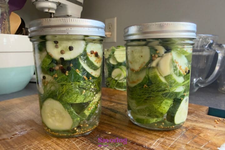 Two glass jars filled with sliced cucumbers, dill, and spices, sitting on a wooden surface, prepared for pickling.