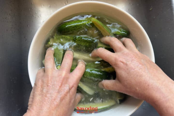 Two hands washing several small cucumbers in a white bowl filled with water, placed in a metal sink.