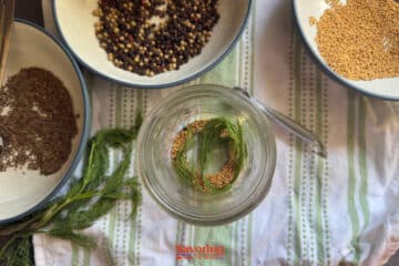 A glass jar of dill pickles with dill and mustard seeds sits on a striped cloth, surrounded by bowls of dried spices and a spoon.