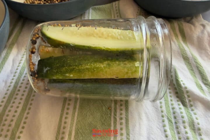 Three cucumber spears and pickling spices in a mason jar, lying on its side on a striped towel.