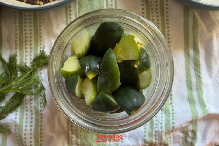 Top-down view of a glass jar filled with cut pickles, placed on a white and green striped cloth with fresh dill visible nearby.