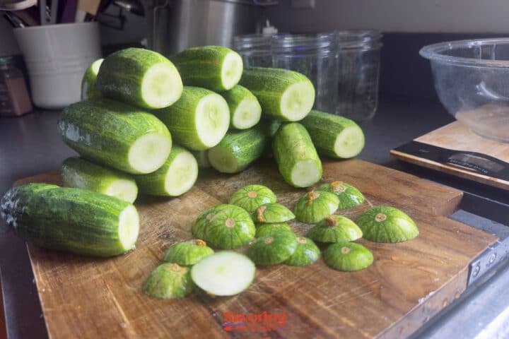A pile of cut cucumbers and sliced stem ends are arranged on a wooden cutting board in a kitchen with jars and utensils in the background, ready to be transformed into crisp dill pickles.