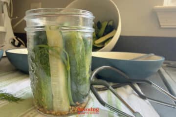 A glass jar filled with cucumber spears, dill, and spices is on a counter next to metal tongs and a bowl of more cucumbers.