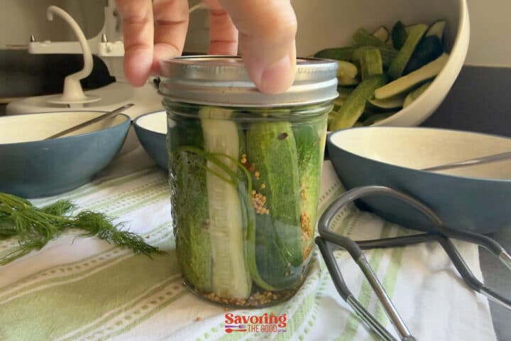 A hand holds a mason jar filled with pickling cucumbers, dill, and spices. Sliced cucumbers, bowls, and kitchen tools are on the counter in the background.