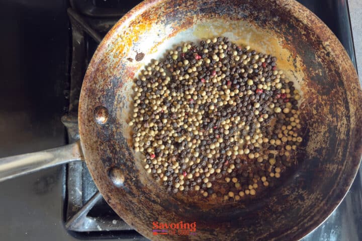 A metal pan with whole peppercorns being toasted on a stovetop burner.