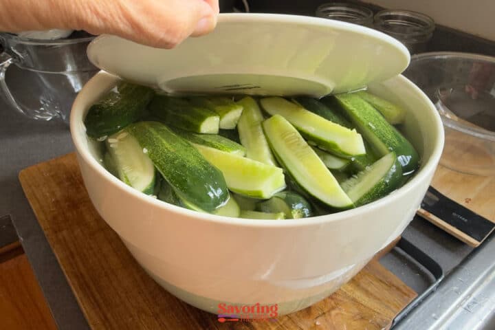 A hand lifts a plate covering sliced cucumbers soaking in water in a large white bowl on a wooden countertop.