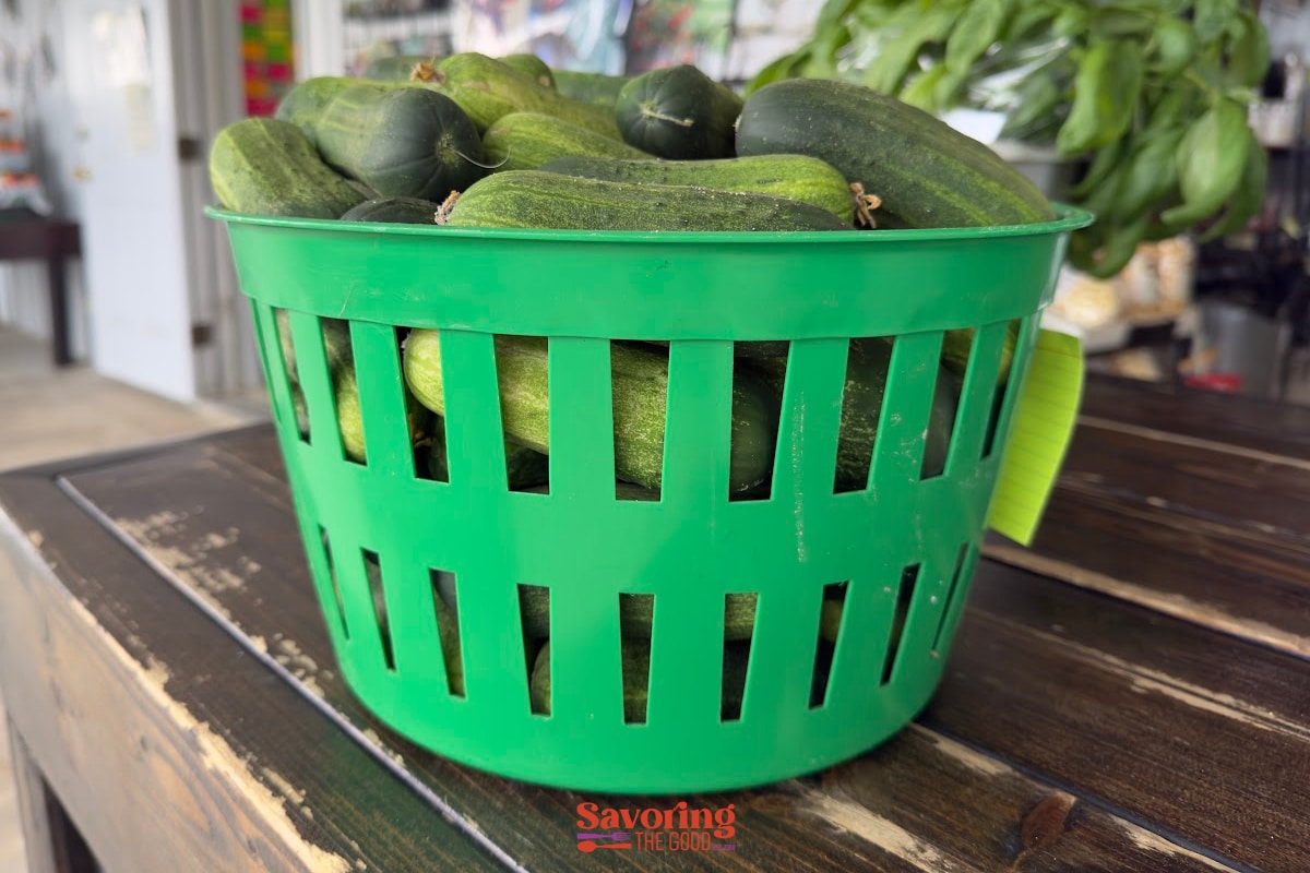 A green plastic basket filled with fresh cucumbers sits on a wooden table.