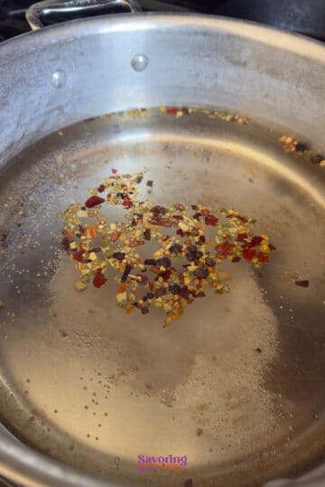 Stainless steel pan with oil heating and red pepper flakes scattered in the center.