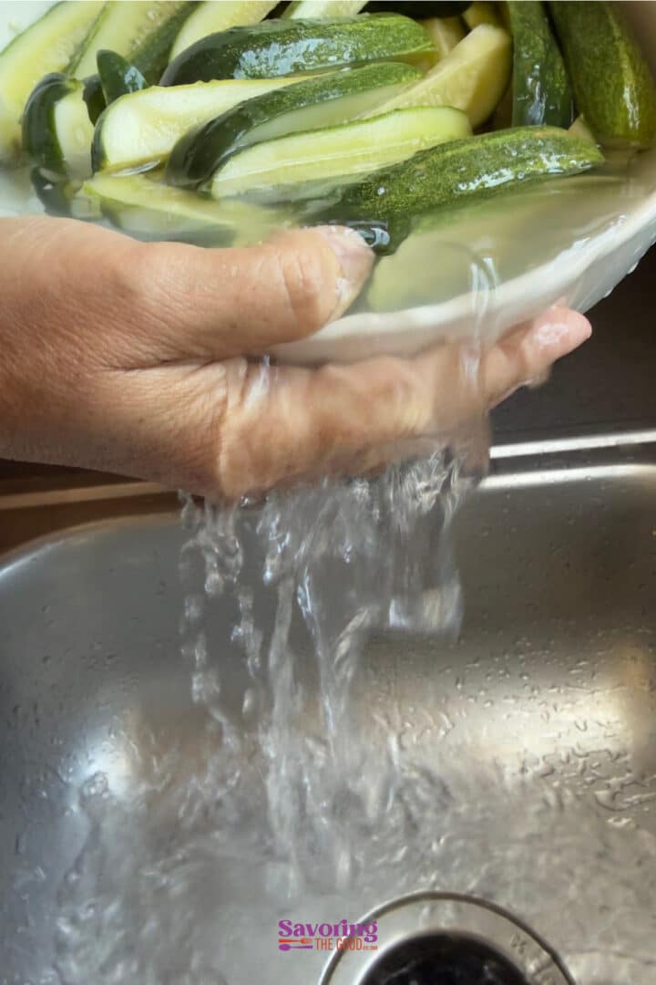 A hand holds a bowl of sliced cucumbers draining under with water overflowing into a sink below.