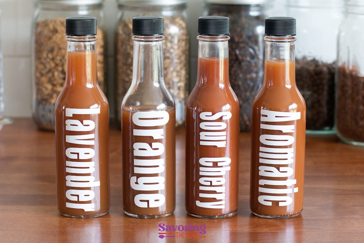 Four glass bottles with brown liquid, labeled Lavender, Orange, Sour Cherry, and Aromatic, are lined up on a wooden counter with jars of ingredients in the background.