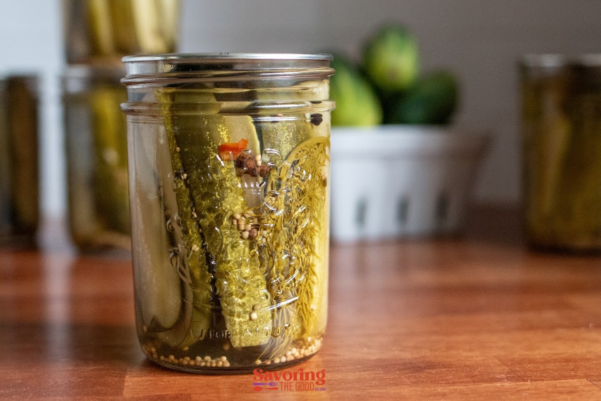 A glass jar of homemade dill pickles with spices sits on a wooden surface, with cucumbers and more jars blurred in the background.