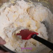 A hand mixes flour and other baking ingredients in a metal bowl with a red spatula.
