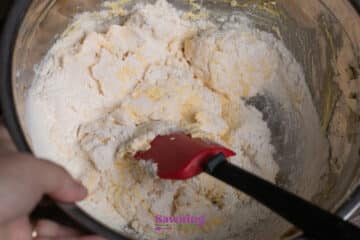 A hand mixes flour and other baking ingredients in a metal bowl with a red spatula.