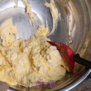 A red spatula sits in a metal bowl containing a yellow, creamy mixture, partially scraped from the sides.