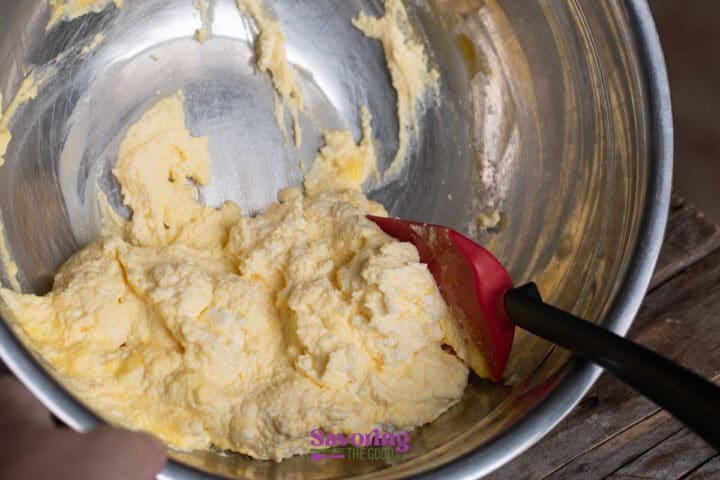 A red spatula sits in a metal bowl containing a yellow, creamy mixture, partially scraped from the sides.