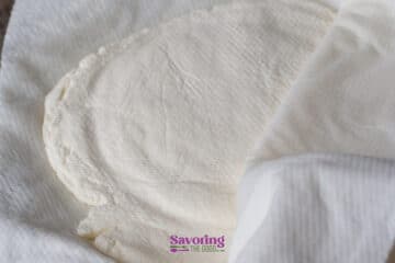 A close-up of homemade ricotta cheese wrapped in a white cloth, partially uncovered, resting on a flat surface.