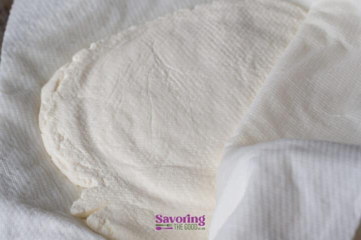 A close-up of homemade ricotta cheese wrapped in a white cloth, partially uncovered, resting on a flat surface.