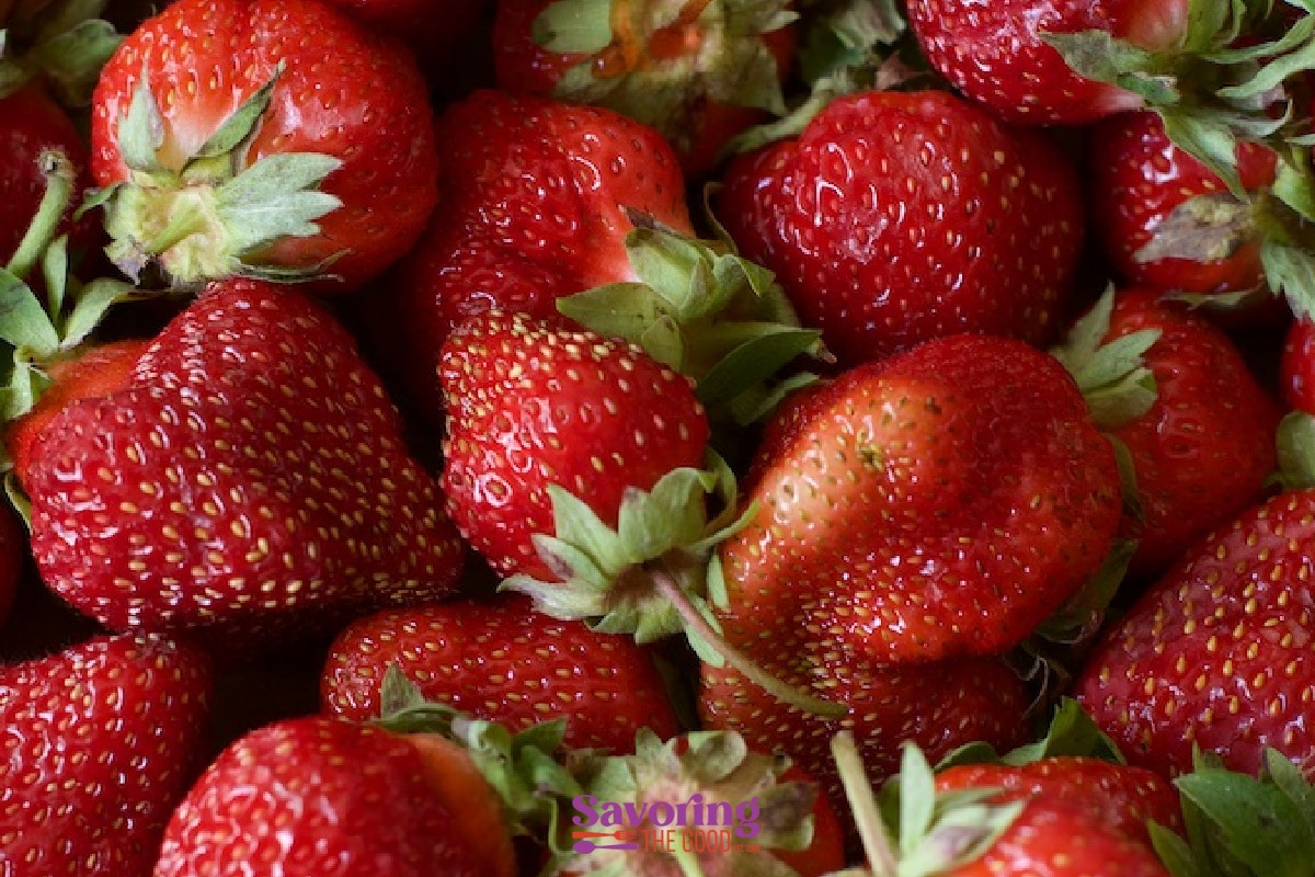 Close-up of a pile of ripe red strawberries with green leaves, showing their textured surfaces and natural variations in size and shape.