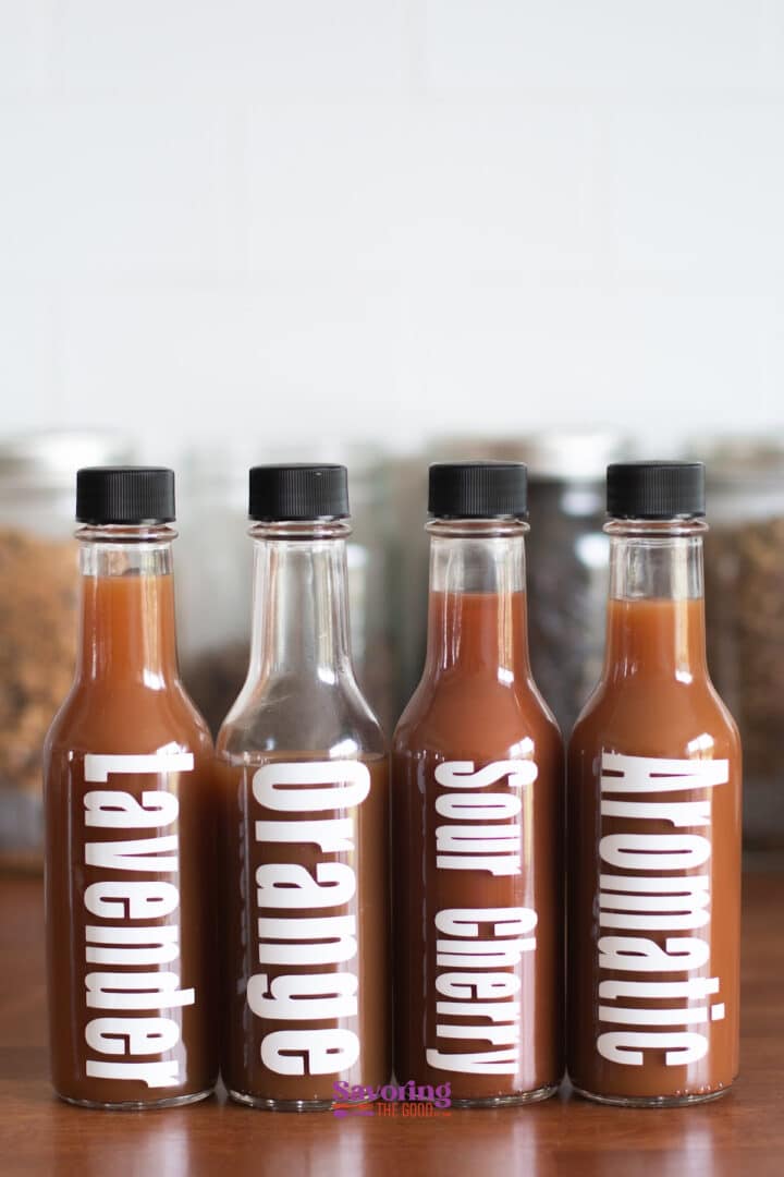 Four bottles labeled Lavender, Orange, Sour Cherry, and Aromatic are filled with reddish-brown liquid and have black caps, set against a blurred background of jars—perfect for anyone eager to learn how to make bitters at home.