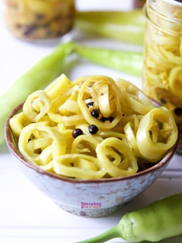 A bowl of sliced pickled banana peppers with visible peppercorns, surrounded by fresh banana peppers and jars of more pickled peppers in the background.