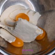 A metal bowl containing ricotta cheese and three raw egg yolks, with a hand holding the rim of the bowl.