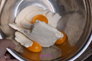 A metal bowl containing ricotta cheese and three raw egg yolks, with a hand holding the rim of the bowl.