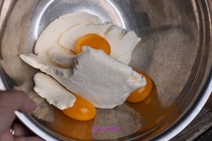 A metal bowl containing ricotta cheese and three raw egg yolks, with a hand holding the rim of the bowl.