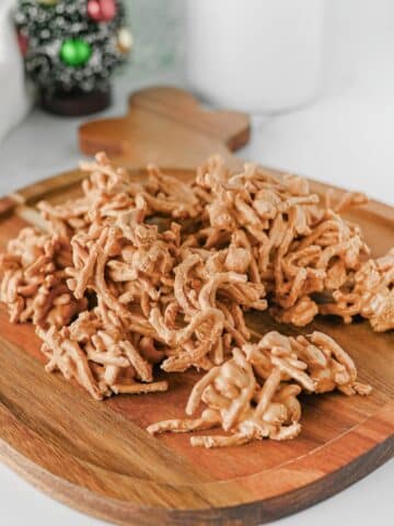 A pile of Butterscotch Haystacks sits on a wooden board, with a blurred Christmas tree decoration in the background.