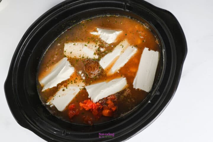 A slow cooker filled with broth, diced tomatoes, seasoning, and several slices of cream cheese on top, ready to be cooked.