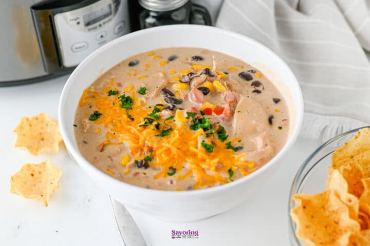 A bowl of creamy chicken soup with black beans, corn, shredded cheese, and herbs-reminiscent of Crockpot cream cheese chicken chili-sits next to tortilla chips with a slow cooker in the background.