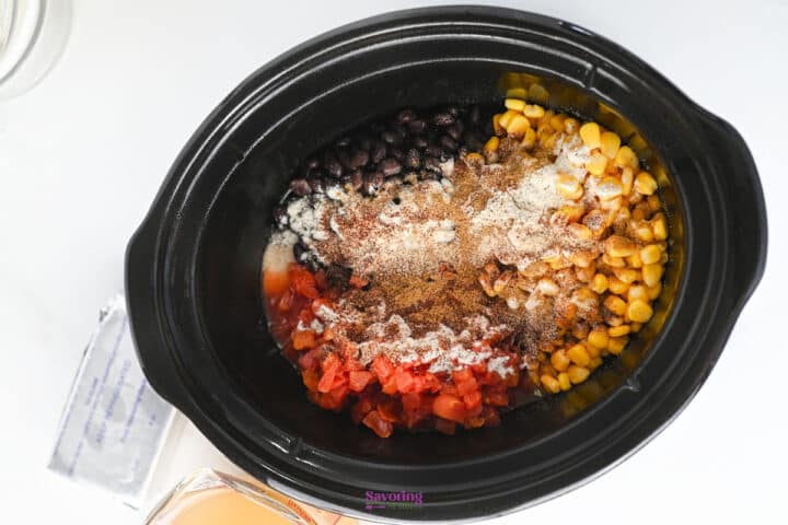 A slow cooker filled with black beans, corn, diced tomatoes, seasonings, and powdered ingredients, ready to be mixed and cooked.