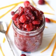 A glass jar filled with homemade Cranberry Apple Chutney sits on a white surface, surrounded by fresh cranberries, orange peel strips, and a spoon.