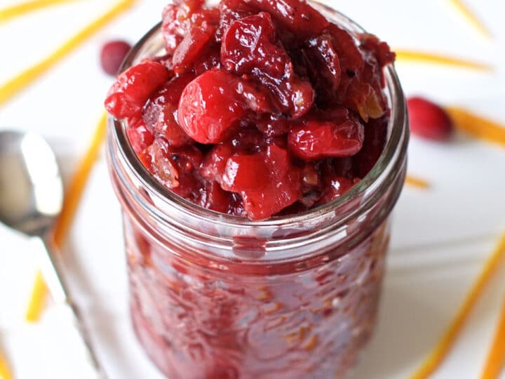 A glass jar filled with homemade Cranberry Apple Chutney sits on a white surface, surrounded by fresh cranberries, orange peel strips, and a spoon.