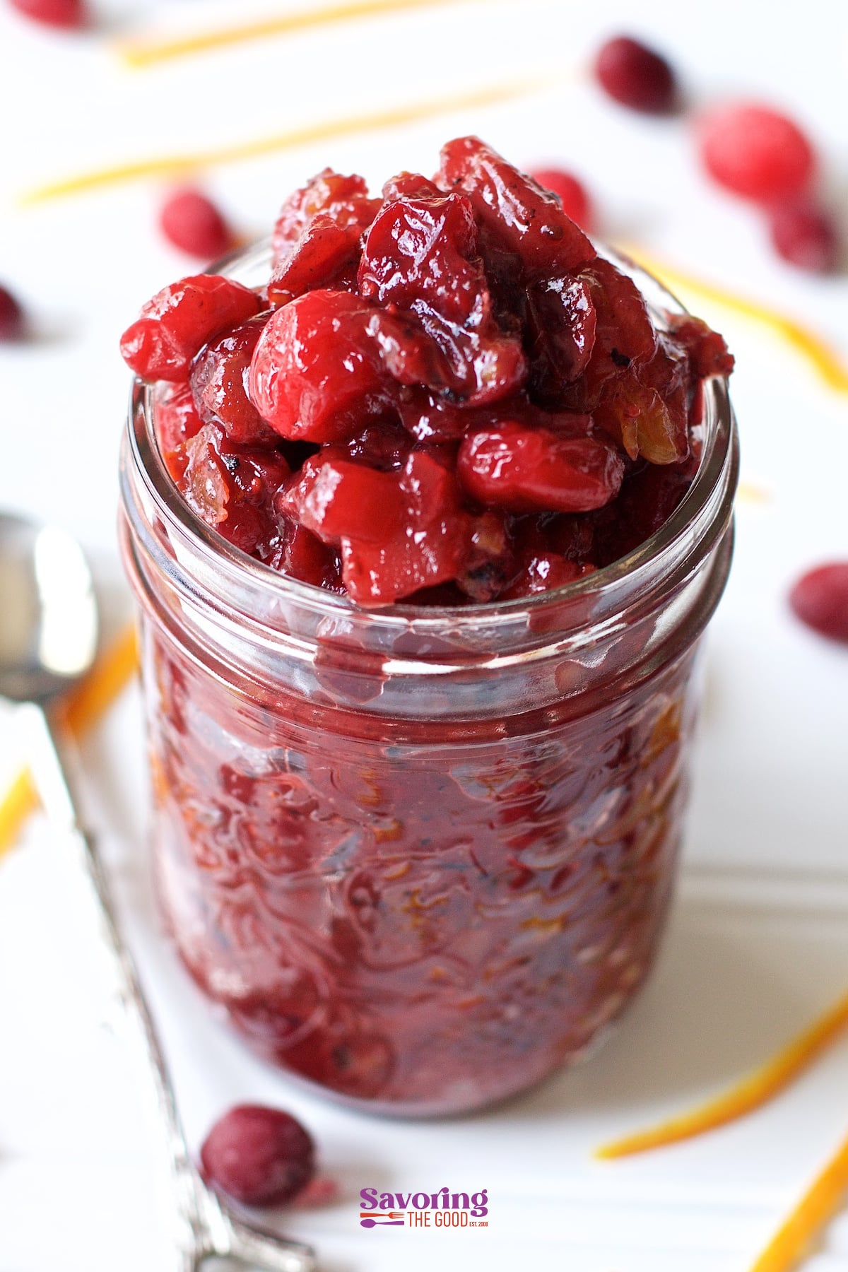 Perfect for your Thanksgiving table my recipe for cranberry apple chutney also has freshly grated ginger, brown sugar, orange zest and an optional kick of heat. By choosing to use fresh cranberries or frozen cranberries you will be making this easy cranberry chutney to go along with your charcuterie and cheese board beyond Thanksgiving. #chutney #thanksgivingsidedish #thanksgiving #cranberrychutney #easychutney #Thanksgivingside #cranberry A glass jar filled with cranberry sauce sits on a white surface, surrounded by loose cranberries and orange zest, with a spoon beside it-an inviting scene reminiscent of homemade Cranberry Apple Chutney.