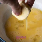 A hand dips a dough ball into a bowl of beaten eggs, preparing it for coating.
