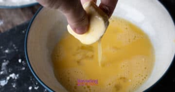 A hand dips a dough ball into a bowl of beaten eggs, preparing it for coating.