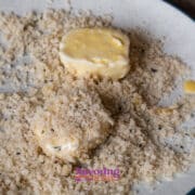 Two coated slices goat cheese are partially covered with breadcrumbs on a white plate. A red container is visible in the background on a wooden surface.