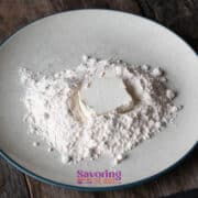 A mound of flour with a square of butter in the center sits on a round, white plate placed on a rustic wooden surface-perfect ingredients for preparing crispy fried goat cheese.