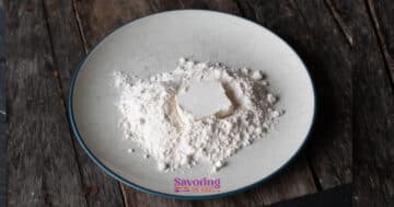 A mound of flour with a square of butter in the center sits on a round, white plate placed on a rustic wooden surface—perfect ingredients for preparing crispy fried goat cheese.
