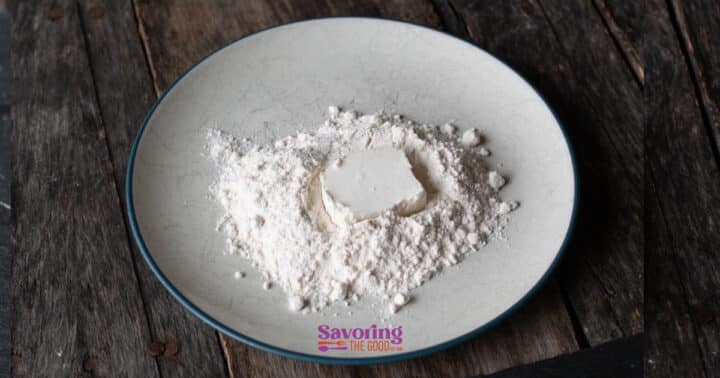 A mound of flour with a square of butter in the center sits on a round, white plate placed on a rustic wooden surface-perfect ingredients for preparing crispy fried goat cheese.